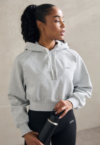 Felpa grigia chiara a maniche corte con cappuccio regolabile e polsini a coste, abbinata a leggings neri. La persona tiene in mano una bottiglia d'acqua nera.