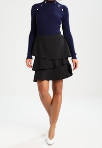 Kvinna iklädd en marinblå ribbad tröja med guldfärgade knappar och en lager-på-lager svart minikjol, kombinerat med vita pumps med spetsiga tår, stående framför en vit bakgrund.