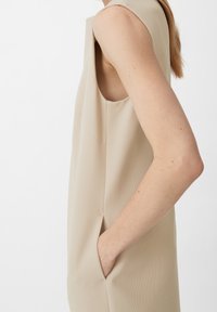 Vestido sin mangas beige con una tela texturizada, que cuenta con bolsillos laterales y un silueta limpia y moderna. Sin cierres visibles ni patrones.
