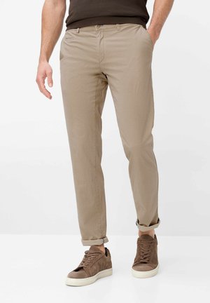 Mand iført beige slim-fit bukser rullet op ved anklerne med brune ruskindssneakers og en mørk skjorte, stående mod en hvid baggrund.