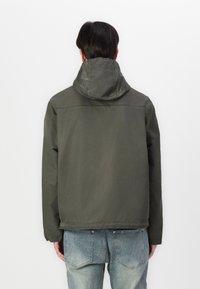 Olivengrønn hettejakke med en avslappet passform, med sømdetaljer og en enkel bakdesign. Brukt med lyseblå jeans.