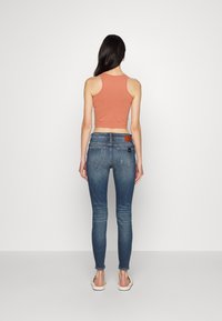Top corto de color naranja claro con tirantes estrechos, combinado con jeans skinny de color azul oscuro. Los jeans presentan un desvanecimiento sutil y un parche en la parte posterior.