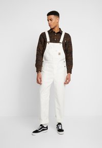 Salopette blanche en coton avec bretelles réglables et poche avant. Associée à une chemise à motifs marron et à des baskets noires.