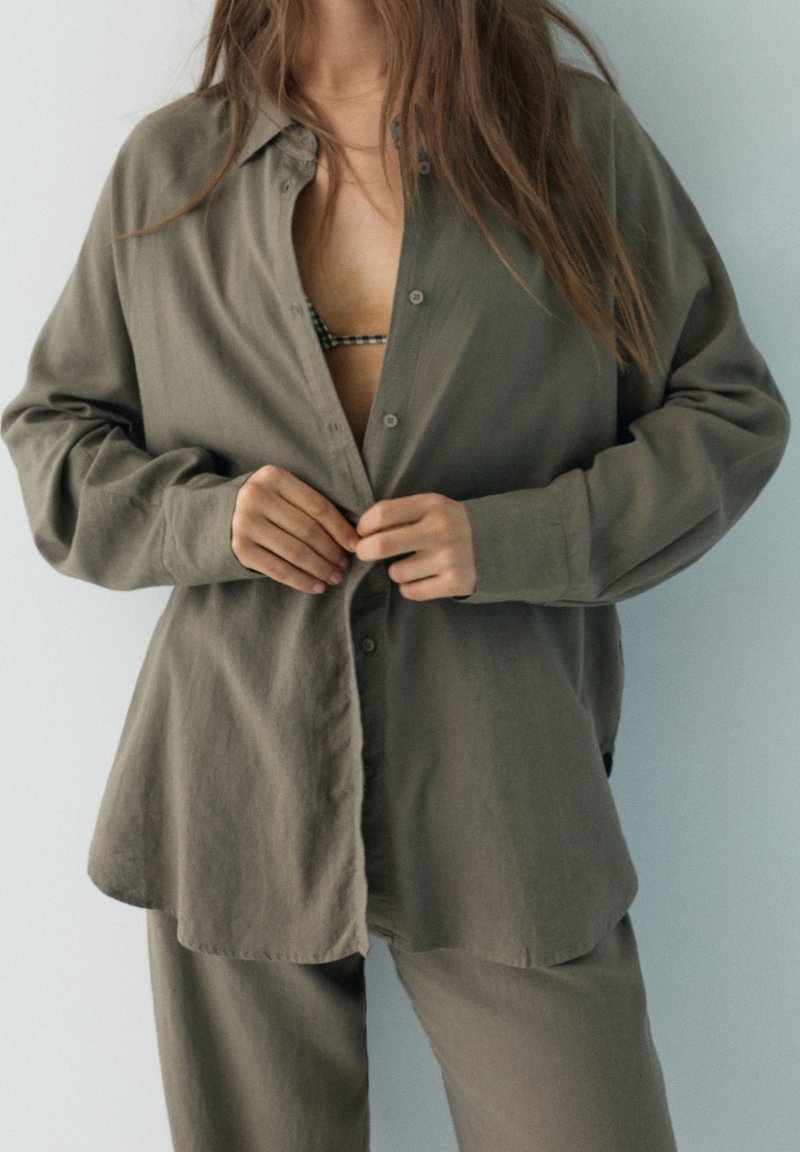 Femme aux longs cheveux boutonnant une chemise ample vert olive par-dessus un pantalon assorti et un soutien-gorge à carreaux visible en dessous.