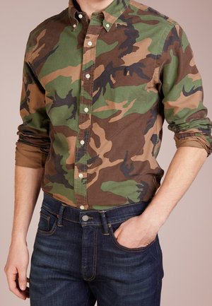Man draagt een groenbruine camouflage blouse met knopen, met opgerolde mouwen, ingestopt in een donkerblauwe spijkerbroek, met één hand in de zak.