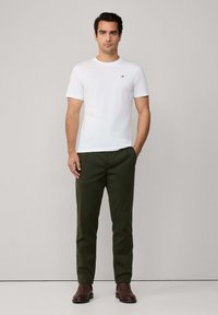 T-shirt bianco a maniche corte con un piccolo logo nero, abbinato a pantaloni verdi e stivali alla caviglia marroni. Design semplice e moderno, colori neutri.