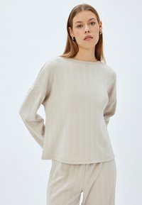 Vaaleanbeige neulottu neulepusero, jossa on ribbikuviointi, pitkät hihat ja pyöreä kaula-aukko. Kankaan pinnalla on hienovaraisia pystysuoria kuvioita.