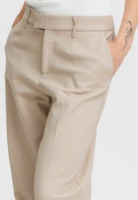 Pantalons beige en tissu lisse et texturé. Comprend des plis sur le devant, des poches latérales et une coupe ajustée. Taille mi-haute avec passants de ceinture.