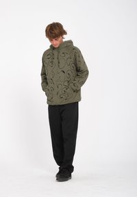 Felpa verde oliva con motivi neri a spirale, tasca frontale e cappuccio con cordino, abbinata a pantaloni neri e scarpe scure.