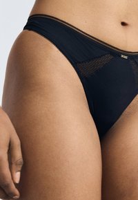 Zwarte bikini slip met mesh-details aan de voorkant, elastische tailleband en een klein merklabel. Glad materiaal met een subtiele glans.