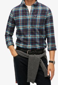 Geruite flanellen shirt in navy, teal en bruin met knoopsluiting. Grijze trui om de taille geknoopt, zwarte riem, spijkerbroek en horloge.