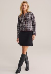 Cardigan dengan pola houndstooth ungu dan putih, leher bulat, tombol emas, dipadukan dengan rok hitam dan sepatu bot hitam setinggi lutut.