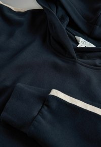 Navy blue hoodie made of cotton blend fabric, featuring a hood and cream accent stripe on the sleeves. Tag visible with size information.