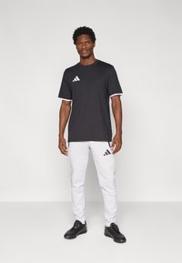 Μαύρο αθλητικό μπλουζάκι με λευκές λεπτομέρειες, με το λογότυπο της Adidas. Συνδυασμένο με λευκό παντελόνι και μαύρα παπούτσια, παρουσιάζοντας μια αθλητική εμφάνιση.