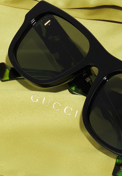 Črne oversize sončna očala z zelenimi detajli na ročkih, postavljena na rumenem blagu, ki prikazuje logotip "GUCCI" v kovinskem tisku.