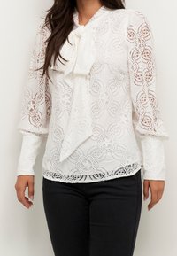 Vit spetsblus med en rosett vid kragen, långa ärmar med intrikata spettdetaljer och en scalloped fåll. Matcher med mörka jeans.