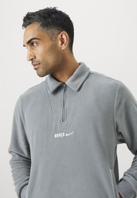 Γκρι φλις πουλόβερ με μισό φερμουάρ και γιακά. Διαθέτει κεντημένο το "BARÇA" και το λογότυπο της Nike σε λευκό χρώμα. Μαλακή υφή, χαλαρή εφαρμογή.