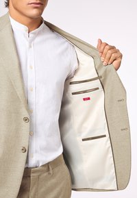 Helle beige Anzugjacke mit einer glatten, texturierten Außenseite. Das Innenfutter ist aus hellfarbigem Satin und hat Taschen. Trägt man über einem weißen Hemd mit Kragen.