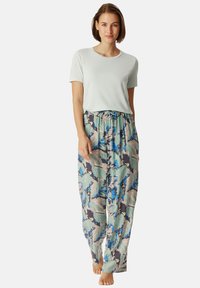 Jeune femme debout pieds nus, portant un haut à manches courtes gris clair et un pantalon floral large et ample.