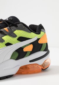 Sko med ett ovandel i mesh i grön och svart, med orange detaljer och en vit sidremsa. Utrustad med en synlig orange luftdämpningsenhet.