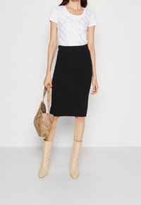 Mujer con una camiseta blanca con estampado, falda negra a la altura de la rodilla, botas beige hasta la mitad de la pantorrilla, sosteniendo un bolso beige con un estampado y asa de hombro.