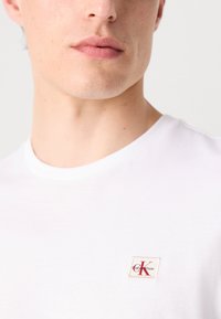 Lähikuva henkilöstä, joka käyttää valkoista paitaa, jossa on pieni punainen ja musta Calvin Klein -logoplässi rinnassa.