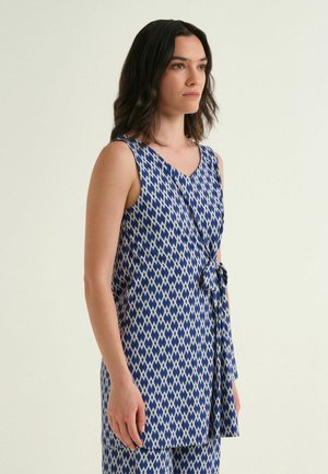 Femme aux cheveux foncés mi-longs, portant une tunique sans manches à motif géométrique bleu et blanc avec un nœud sur le côté, debout devant un fond uni.