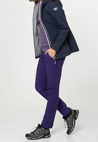 Giacca blu navy impermeabile con fodera grigia, abbinata a pantaloni impermeabili viola e scarpe da trekking nere con suole texture.