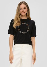 Zwarte katoenen T-shirt met een oversized pasvorm. Ronde witte en oranje tekstgrafiek gedrukt aan de voorkant. Korte mouwen en ronde hals.