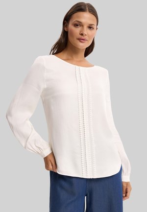 Kobieta o długich brązowych włosach, ubrana w białą bluzkę z długim rękawem, ozdobioną pionowym, fakturowanym zdobieniem, oraz niebieskie spodnie, stojąca z jedną ręką na biodrze.