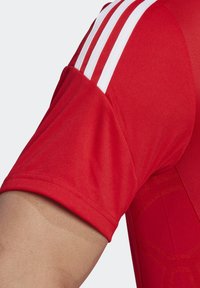 Camisa atlética roja con mangas cortas, tres rayas blancas en el hombro y un patrón texturizado en la tela.