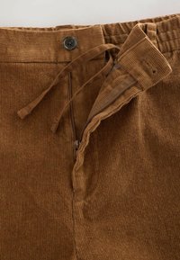 Pantalon en velours côtelé marron avec fermeture éclair et cordon de serrage. Texture rainurée notable et une seule fermeture à bouton à la taille.