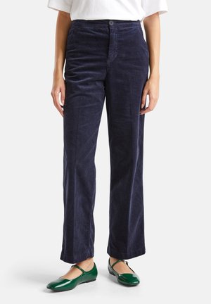 Vrouw in marineblauwe corduroy rechte broek met groene Mary Jane schoenen en een wit shirt, staande tegen een witte achtergrond.