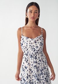 Robe en satin avec un motif à pois bleu marine et blanc, décolleté en cowl, bretelles spaghetti réglables et détail de taille nouée.