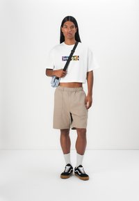 Vit kortärmad t-shirt med färggård "Converse"-logotyp, beige shorts med fickor, grå strumpor, svarta sneakers med vita detaljer, minimalistisk stil.