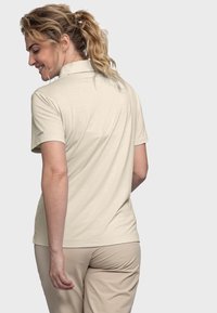Frau mit blonden, lockigen Pferdeschwanz, die ein beiges, kurzärmeliges Poloshirt und passende Hosen trägt, sich mit einem leichten Lächeln abwendet.