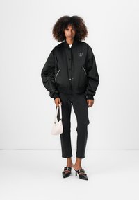 Zwarte satijnen bomberjack met een klein hartlogo, witte piping, gedragen over donkere jeans, gecombineerd met een roze handtas en zwarte hoge instapschoenen.