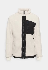 Giacca in pile di colore avorio con accenti neri, dotata di colletto alto, zip frontale e tasca sul petto con zip. Tessuto testurizzato con una finitura morbida.