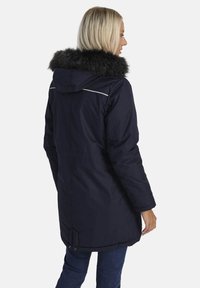 Sieviete, tērpta tumši zilā ziemas mētelī ar kapuci, kas iekšpusē odēta ar melnu mākslīgo vilnu, stāv un ir nedaudz pagriezusies pa labi uz balta fona.