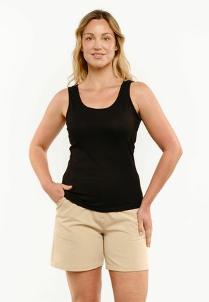 Kvinde står med den ene hånd i lommen på beige shorts, iført en sort ærmeløs top, vendt fremad mod en hvid baggrund.