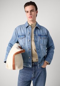 Light blue denim jacket with two chest pockets, button closures, and branded tag. Beige shirt underneath and a cream bag with brown accents.