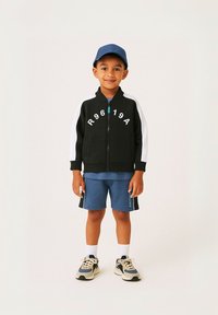 Veste noire zippée avec des manches blanches et des lettres imprimées. T-shirt bleu et shorts. Baskets claires avec des accents sombres et chaussettes blanches.