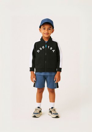 Giacca nera con zip e maniche bianche con lettere stampate. T-shirt blu e pantaloni corti. Sneakers chiare con accenti scuri e calzini bianchi.