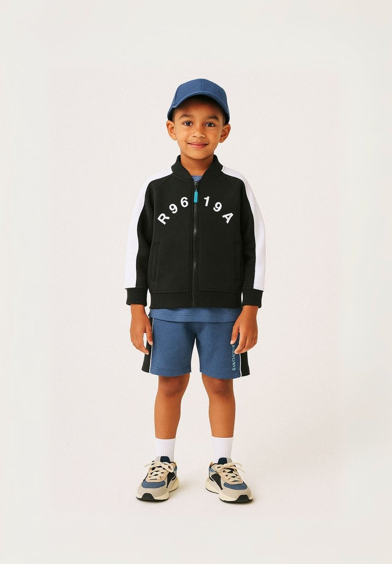 Veste noire zippée avec des manches blanches et des lettres imprimées. T-shirt bleu et shorts. Baskets claires avec des accents sombres et chaussettes blanches.