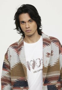 Wool blend cardigan with tribal pattern in grey, brown, and rust colors. Layered over a white t-shirt with printed text and feather design.