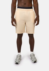 Pantaloni sportivi beige chiari con un design a coste testurizzato, con vita elastica e tasche laterali, abbinati a sneakers grigie e calze bianche.
