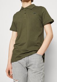 Olivově zelené polo tričko s krátkým rukávem klasického střihu, se třemi knoflíky a malým detailem nášivky na levé hrudi. Spárované se šedými opotřebovanými džíny.
