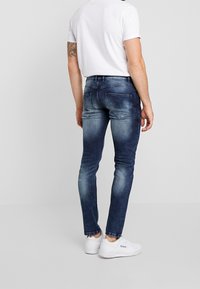 Hombre vestido con jeans ajustados de color azul deslavado, camiseta blanca y zapatillas Reebok blancas, de pie contra un fondo gris liso.