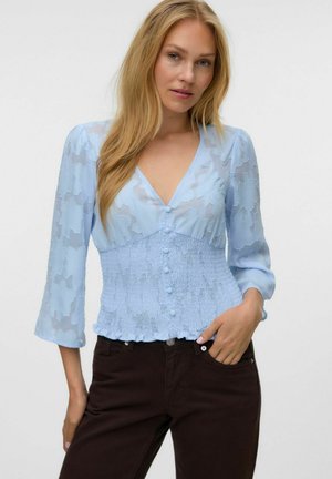 Femme blonde portant un chemisier texturé bleu clair avec des boutons et un pantalon noir, debout avec une main dans la poche devant un fond uni.