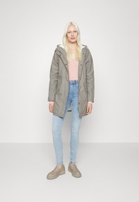 Casaco cinza com capuz por cima de uma blusa rosa; jeans skinny de lavagem clara; sapatos beges texturizados tipo slip-on; modelo a posar sobre um fundo simples.